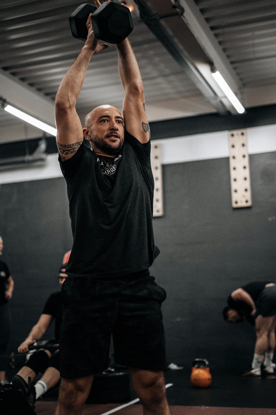 Man with beard lifts dumbbell overhead in gym.